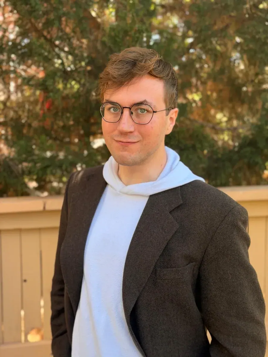 Michał Bodzianowski, smiling, wearing round glasses, a white hoodie and a dark tweed blazer, outdoors