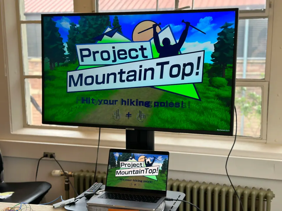 Project Mountaintop title screen on a monitor, with a laptop running the game in front of it